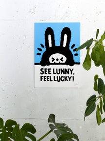 SEE LUNNY, FEEL LUCKY!, TAROART by TAROUT 00029, displayed on a white wall beside a green plant