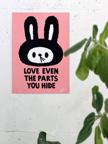 LOVE EVEN THE PARTS YOU HIDE, TAROART by TAROUT 00031, displayed on a white wall beside a green plant