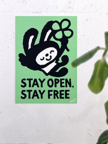 STAY OPEN, STAY FREE, TAROART by TAROUT 00032, displayed on a white wall beside a green plant