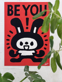 BE YOU!, TAROART by TAROUT 00033, displayed on a white wall beside a green plant